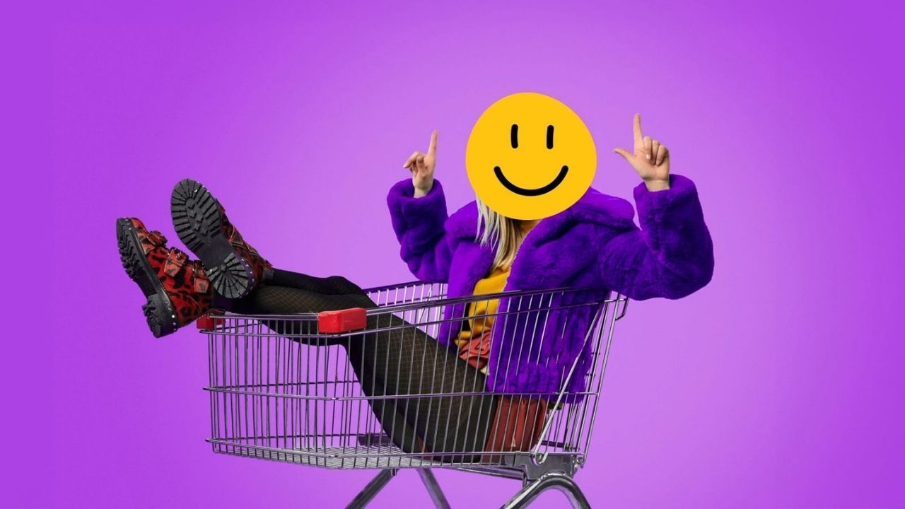 Person wearing a purple coat sitting inside a shopping cart with a yellow smiley face emoji covering their head, pointing fingers upward.